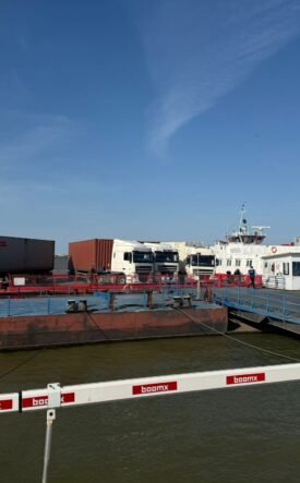 Port scene with a red oil barge, white trucks, and a pier with blue railings under a clear blue sky.