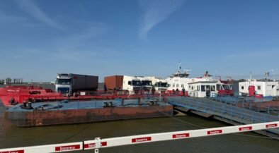 Port scene with a red oil barge, white trucks, and a pier with blue railings under a clear blue sky.