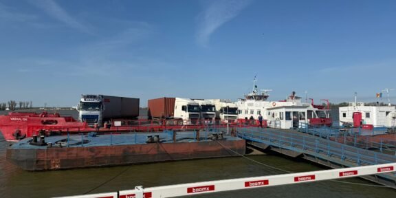Port scene with a red oil barge, white trucks, and a pier with blue railings under a clear blue sky.