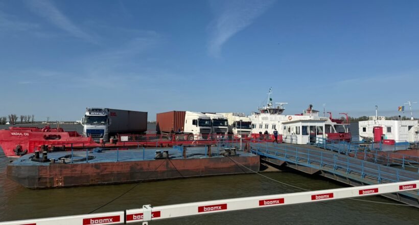 Port scene with a red oil barge, white trucks, and a pier with blue railings under a clear blue sky.