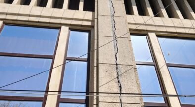 Beige stone building facade with a tall vertical crack running through a central pillar between two windows, under a blue sky, with overhead wires crossing the wall.