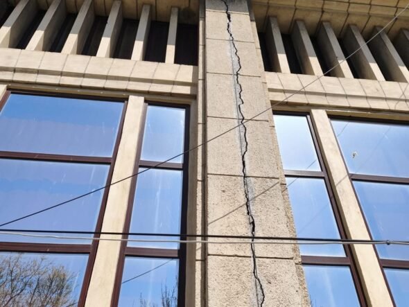 Beige stone building facade with a tall vertical crack running through a central pillar between two windows, under a blue sky, with overhead wires crossing the wall.