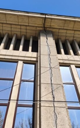 Beige stone building facade with a tall vertical crack running through a central pillar between two windows, under a blue sky, with overhead wires crossing the wall.
