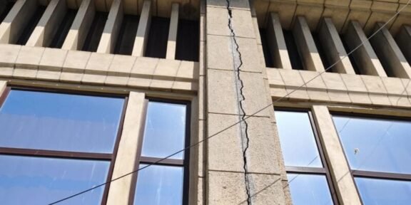 Beige stone building facade with a tall vertical crack running through a central pillar between two windows, under a blue sky, with overhead wires crossing the wall.