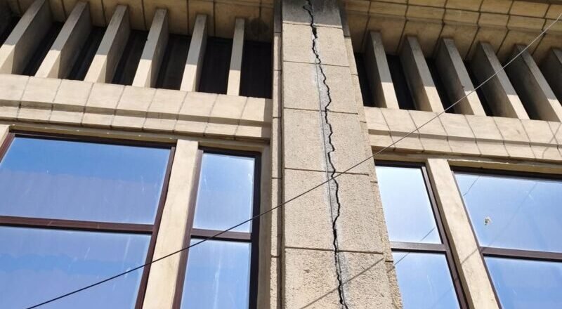 Beige stone building facade with a tall vertical crack running through a central pillar between two windows, under a blue sky, with overhead wires crossing the wall.