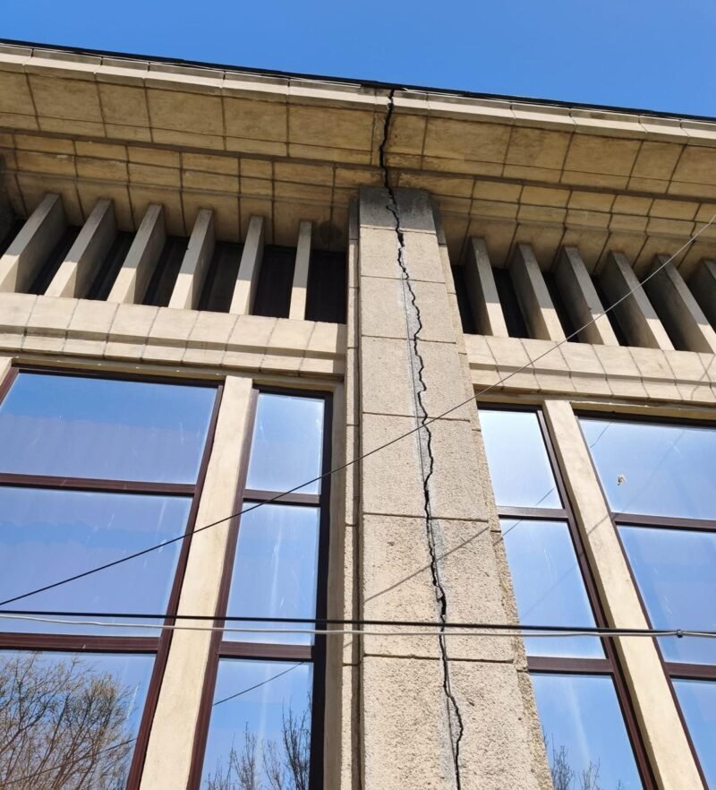 Beige stone building facade with a tall vertical crack running through a central pillar between two windows, under a blue sky, with overhead wires crossing the wall.
