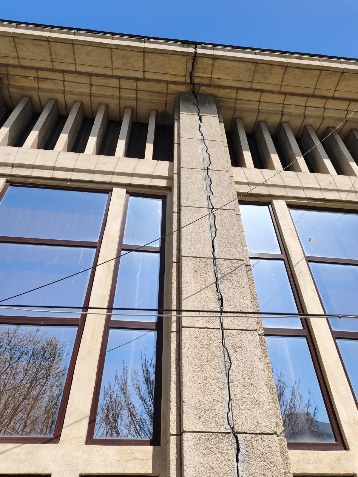 Beige stone building facade with a tall vertical crack running through a central pillar between two windows, under a blue sky, with overhead wires crossing the wall.
