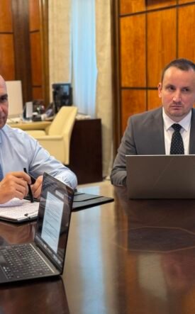 Two businessmen in suits sit at a large conference table with laptops and documents, flags in the background.