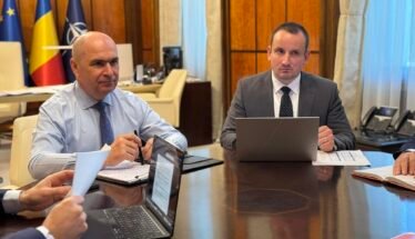 Two businessmen in suits sit at a large conference table with laptops and documents, flags in the background.