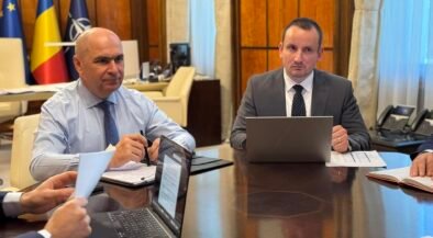 Two businessmen in suits sit at a large conference table with laptops and documents, flags in the background.