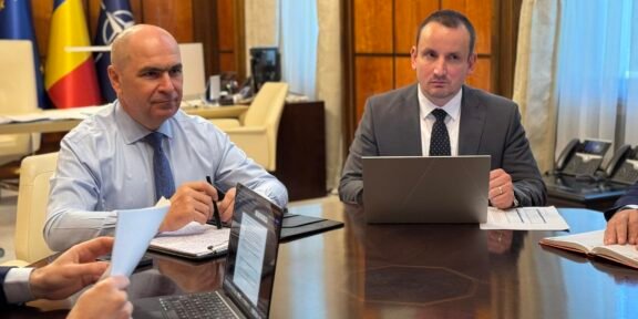 Two businessmen in suits sit at a large conference table with laptops and documents, flags in the background.