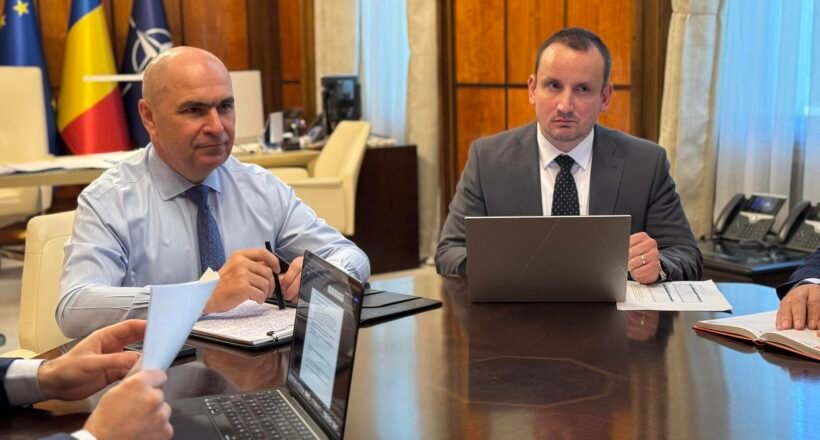 Two businessmen in suits sit at a large conference table with laptops and documents, flags in the background.