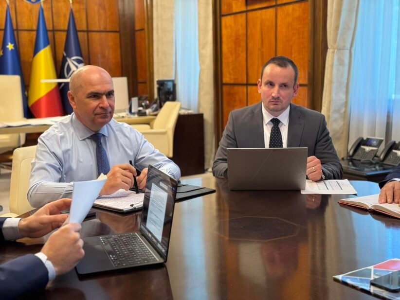 Two businessmen in suits sit at a large conference table with laptops and documents, flags in the background.