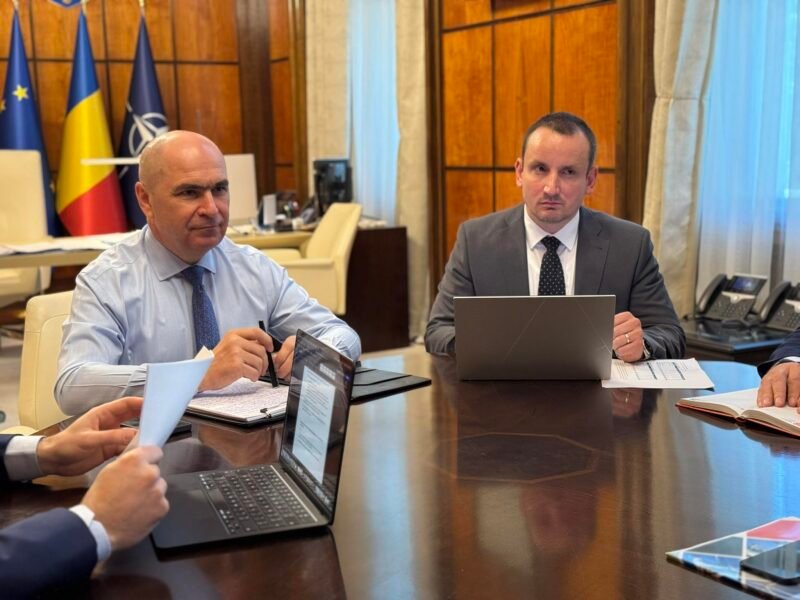 Two businessmen in suits sit at a large conference table with laptops and documents, flags in the background.