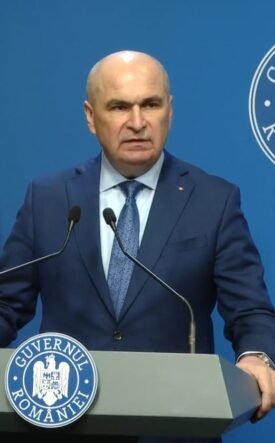 Romanian official giving a press conference at a podium, with national flag and government seal in the background.