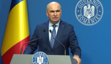 Romanian official giving a press conference at a podium, with national flag and government seal in the background.