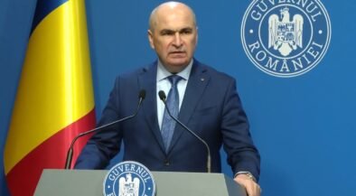 Romanian official giving a press conference at a podium, with national flag and government seal in the background.