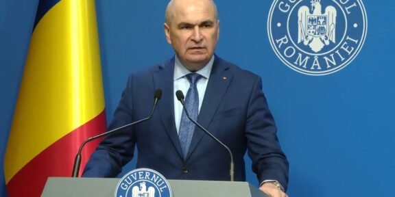 Romanian official giving a press conference at a podium, with national flag and government seal in the background.