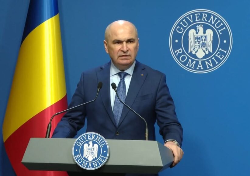 Romanian official giving a press conference at a podium, with national flag and government seal in the background.