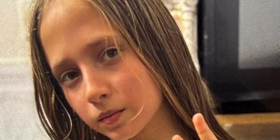 Young girl with long light brown hair making a peace sign, wearing a white shirt with fruit patches indoors.
