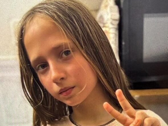 Young girl with long light brown hair making a peace sign, wearing a white shirt with fruit patches indoors.