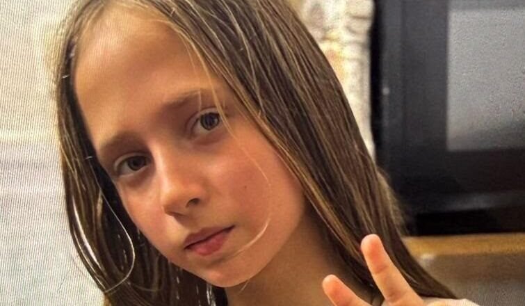 Young girl with long light brown hair making a peace sign, wearing a white shirt with fruit patches indoors.