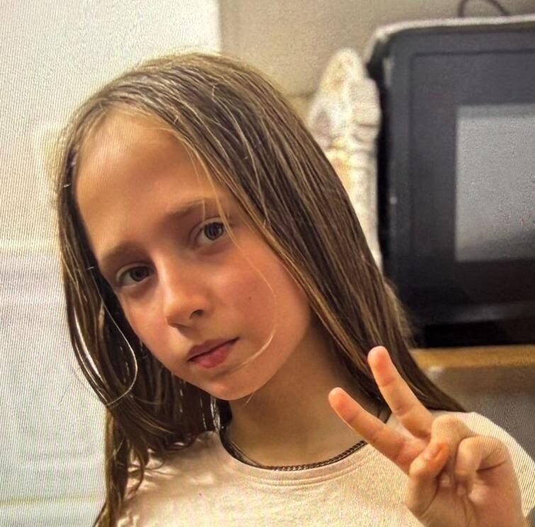Young girl with long light brown hair making a peace sign, wearing a white shirt with fruit patches indoors.
