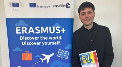 Young man in a black suit stands next to an Erasmus+ banner, holding a colorful diploma.