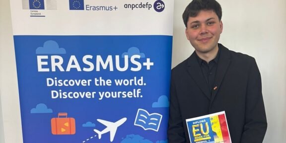 Young man in a black suit stands next to an Erasmus+ banner, holding a colorful diploma.
