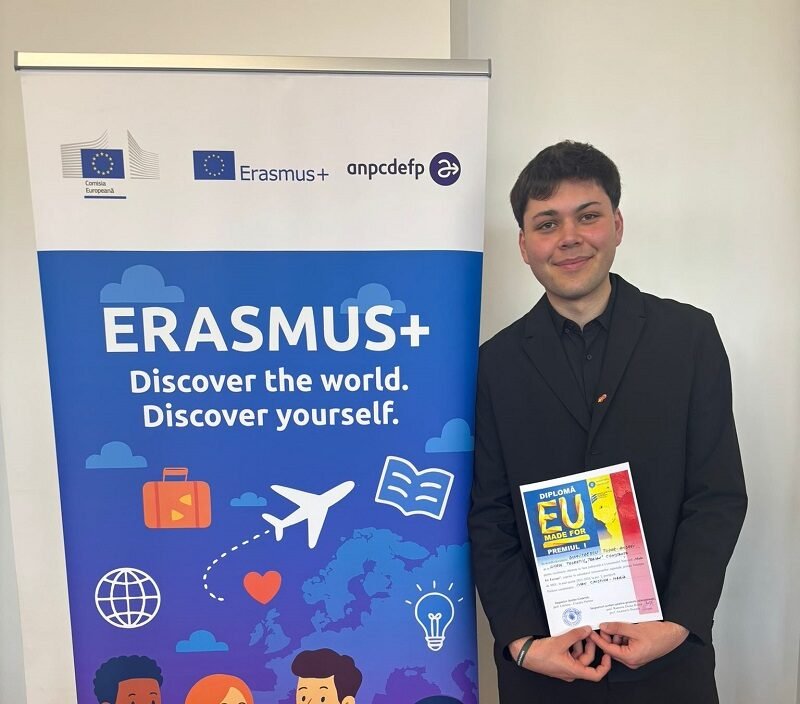 Young man in a black suit stands next to an Erasmus+ banner, holding a colorful diploma.