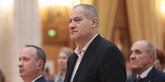 Middle-aged man in a dark suit standing at a formal event, with others in the background.