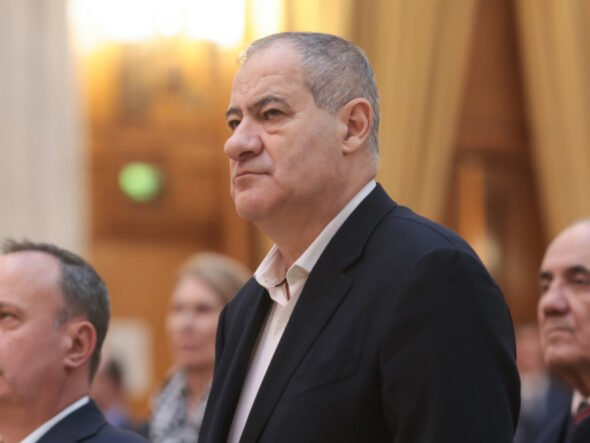 Middle-aged man in a dark suit standing at a formal event, with others in the background.