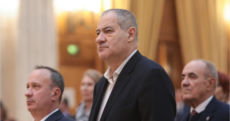 Middle-aged man in a dark suit standing at a formal event, with others in the background.