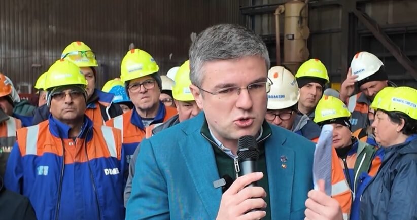Man in a teal jacket speaks into a microphone at a construction site, surrounded by workers wearing yellow hard hats and orange-and-blue safety vests.