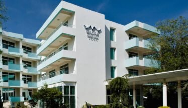 White modern hotel building with multiple floors and turquoise balconies, a glass entrance, and a 'MIRAGE' sign on the façade under a bird logo, set against a clear blue sky.