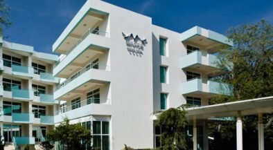 White modern hotel building with multiple floors and turquoise balconies, a glass entrance, and a 'MIRAGE' sign on the façade under a bird logo, set against a clear blue sky.