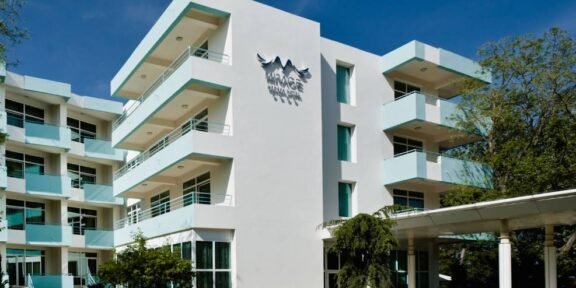 White modern hotel building with multiple floors and turquoise balconies, a glass entrance, and a 'MIRAGE' sign on the façade under a bird logo, set against a clear blue sky.