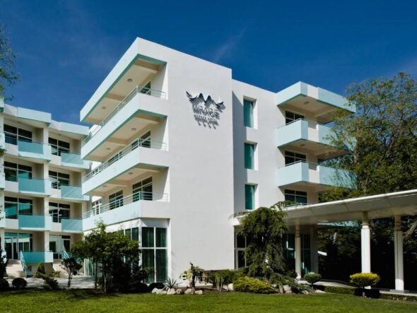 White modern hotel building with multiple floors and turquoise balconies, a glass entrance, and a 'MIRAGE' sign on the façade under a bird logo, set against a clear blue sky.