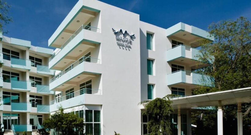 White modern hotel building with multiple floors and turquoise balconies, a glass entrance, and a 'MIRAGE' sign on the façade under a bird logo, set against a clear blue sky.