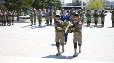 Two soldiers in camouflage carry a wreath toward camera, followed by a crowd of uniformed troops in formation with flags during a ceremony.