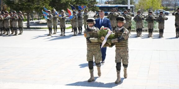 Two soldiers in camouflage carry a wreath toward camera, followed by a crowd of uniformed troops in formation with flags during a ceremony.