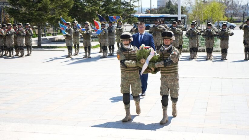 Two soldiers in camouflage carry a wreath toward camera, followed by a crowd of uniformed troops in formation with flags during a ceremony.