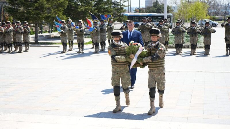 Two soldiers in camouflage carry a wreath toward camera, followed by a crowd of uniformed troops in formation with flags during a ceremony.