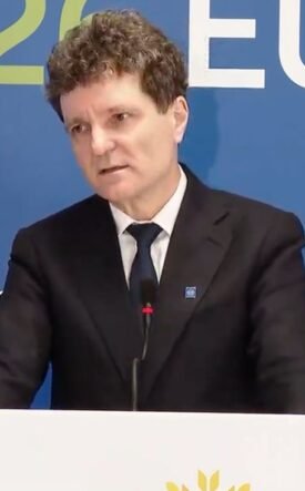 Man in a dark suit giving a speech at a podium, with a blue backdrop that says EU and Cyprus Presidency behind him.