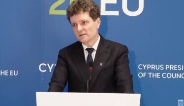 Man in a dark suit giving a speech at a podium, with a blue backdrop that says EU and Cyprus Presidency behind him.