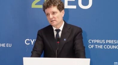 Man in a dark suit giving a speech at a podium, with a blue backdrop that says EU and Cyprus Presidency behind him.