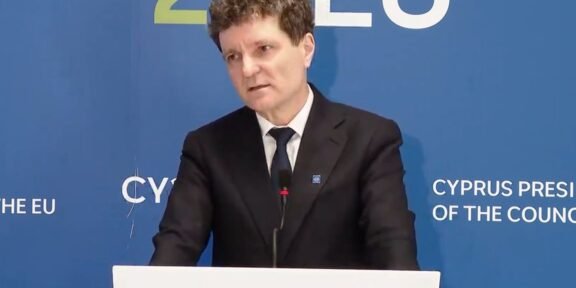 Man in a dark suit giving a speech at a podium, with a blue backdrop that says EU and Cyprus Presidency behind him.