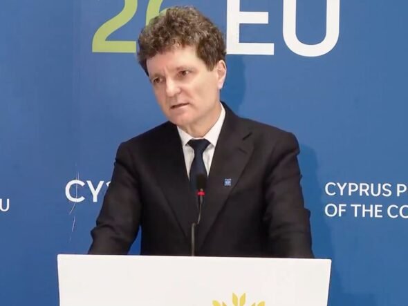 Man in a dark suit giving a speech at a podium, with a blue backdrop that says EU and Cyprus Presidency behind him.
