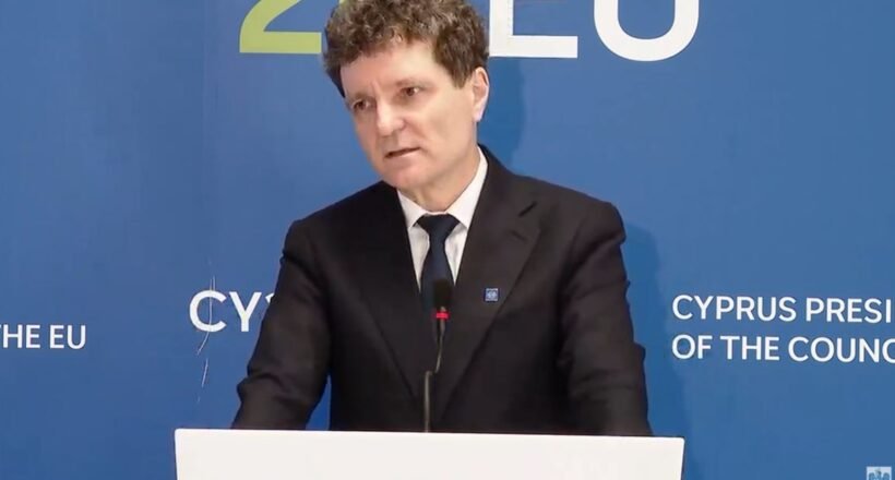 Man in a dark suit giving a speech at a podium, with a blue backdrop that says EU and Cyprus Presidency behind him.