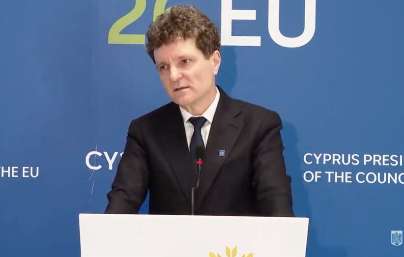 Man in a dark suit giving a speech at a podium, with a blue backdrop that says EU and Cyprus Presidency behind him.